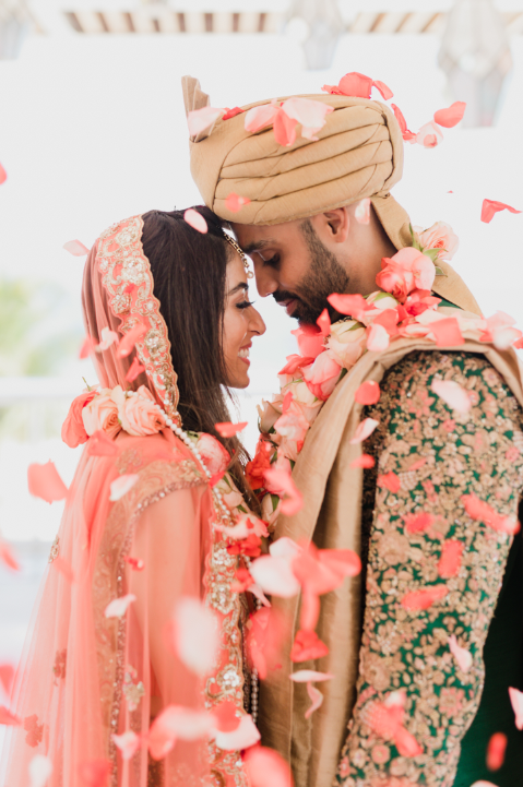 A romantic couple in traditional South Asian wedding attire share an intimate moment amid coral flower petals.