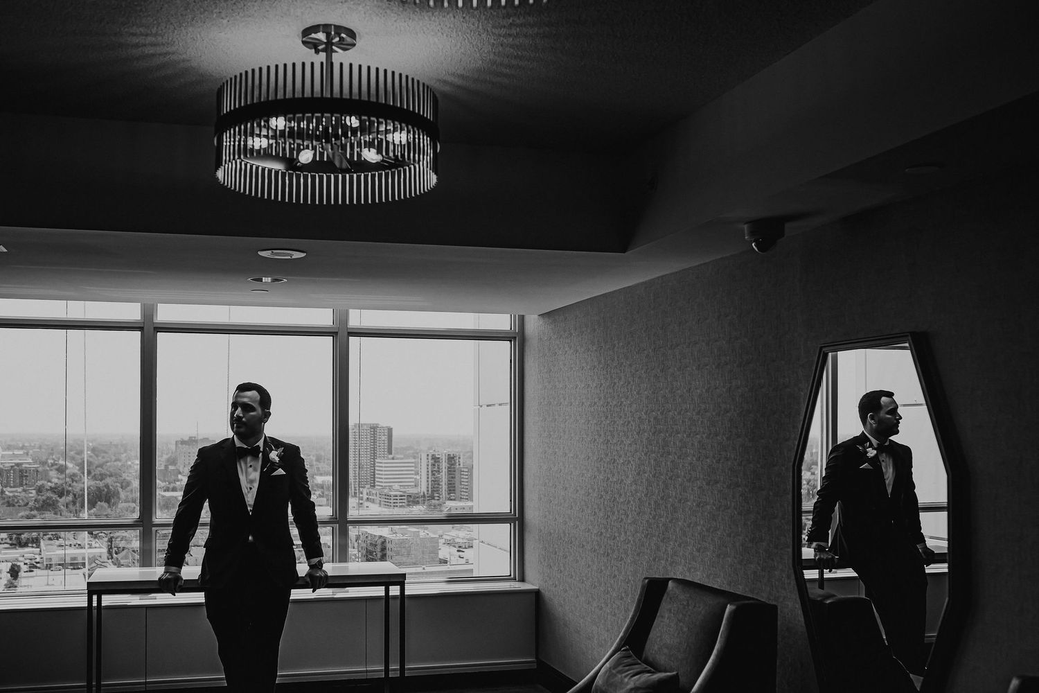 Black and white photo of groom in hotel room with city view and mirror reflection.