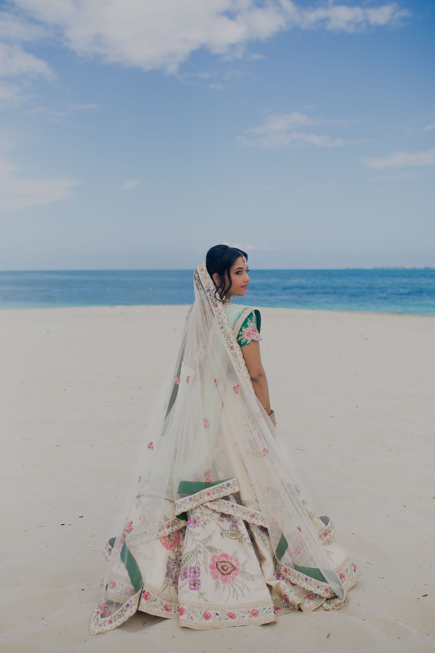 Bride in white floral lehenga poses gracefully on beach with flowing veil.