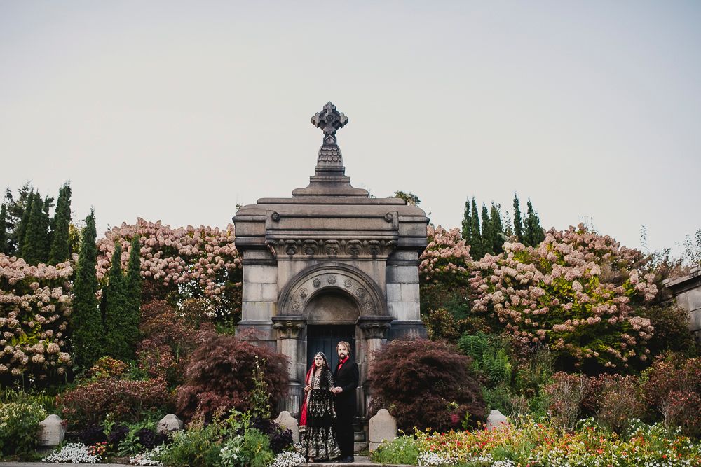 A couple stands at an ornate stone mausoleum entrance surrounded by blooming pink flowers and cypress trees.
