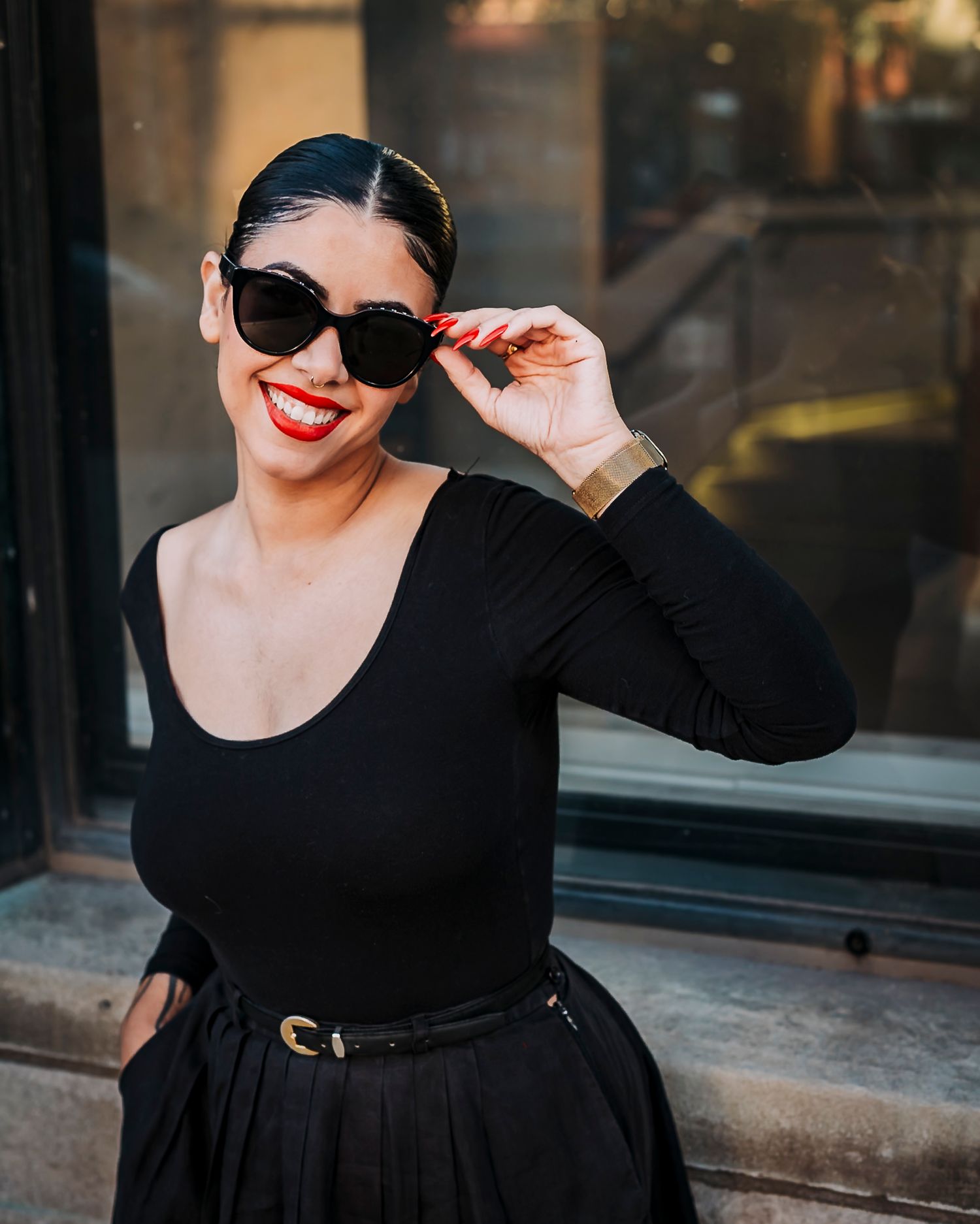 Fashionable woman in black dress and sunglasses smiles confidently at camera.