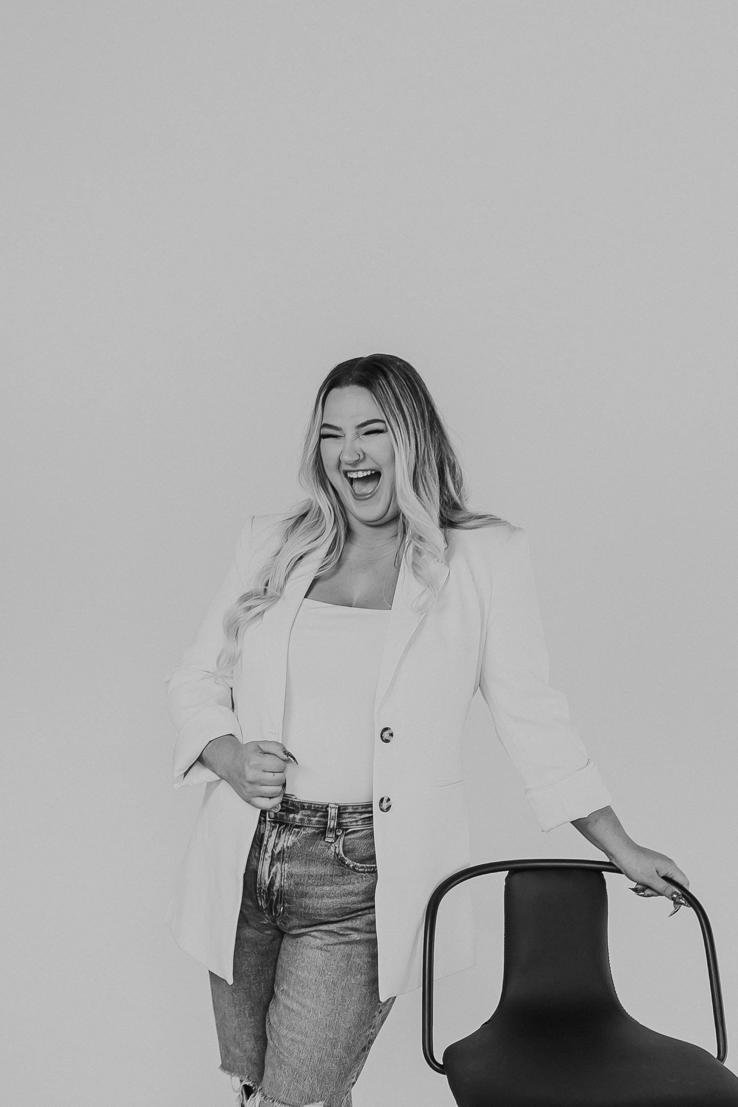 Woman in white blazer and jeans laughing beside a modern chair against a light gray background.