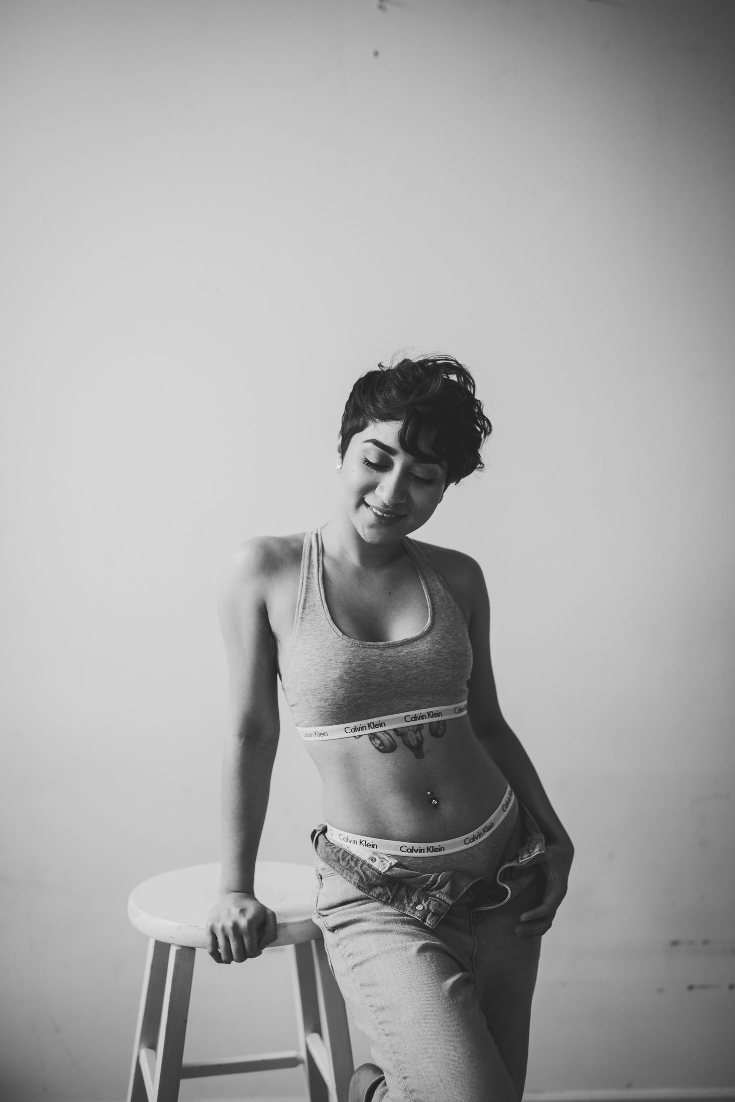 Black and white photo of a young woman in a crop top and jeans posing next to a white stool.