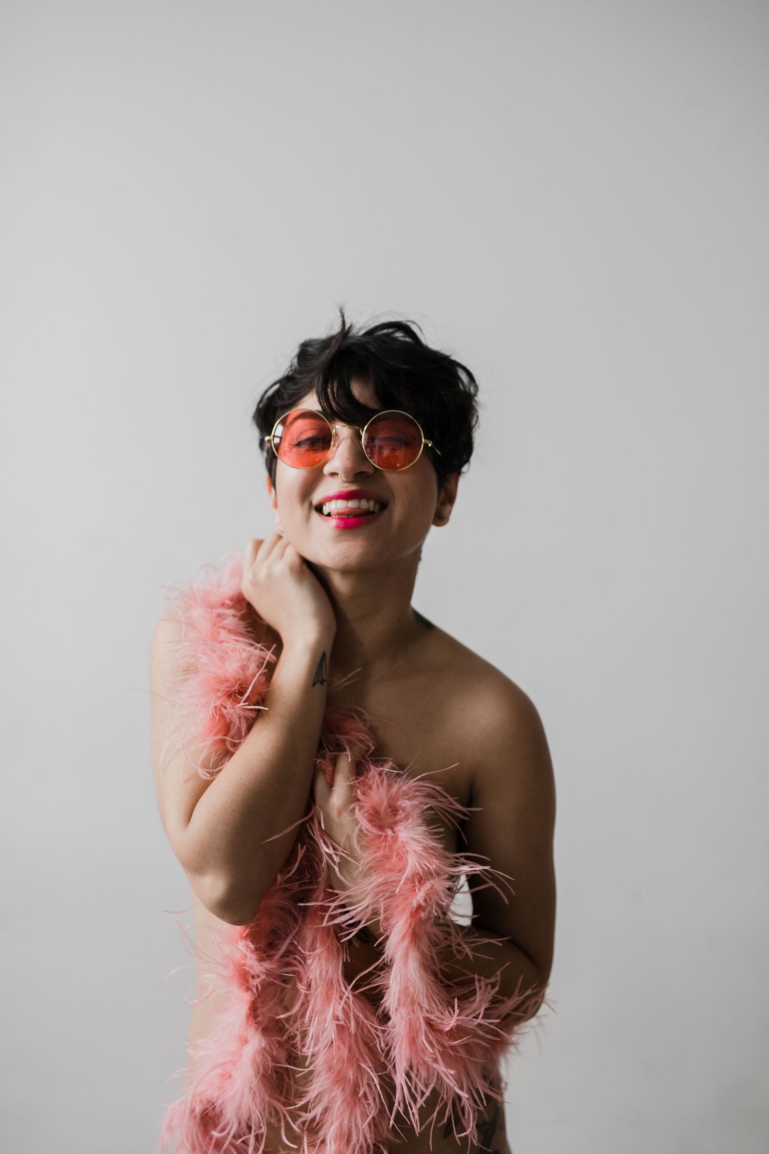 Smiling person wearing pink feather boa and round rose-tinted sunglasses posing against a light gray background.