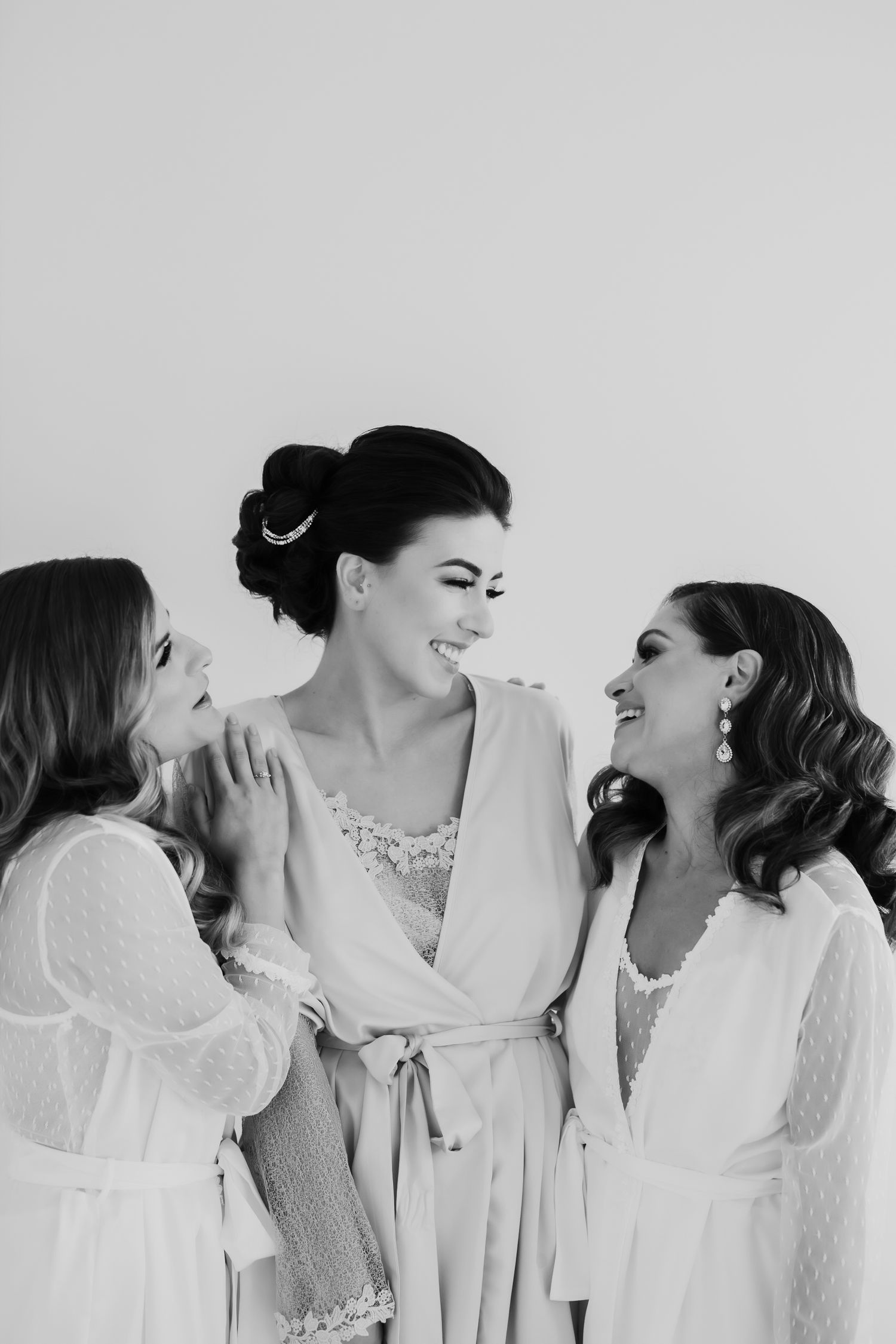 Three women in white robes share joyful laughter during wedding preparation.