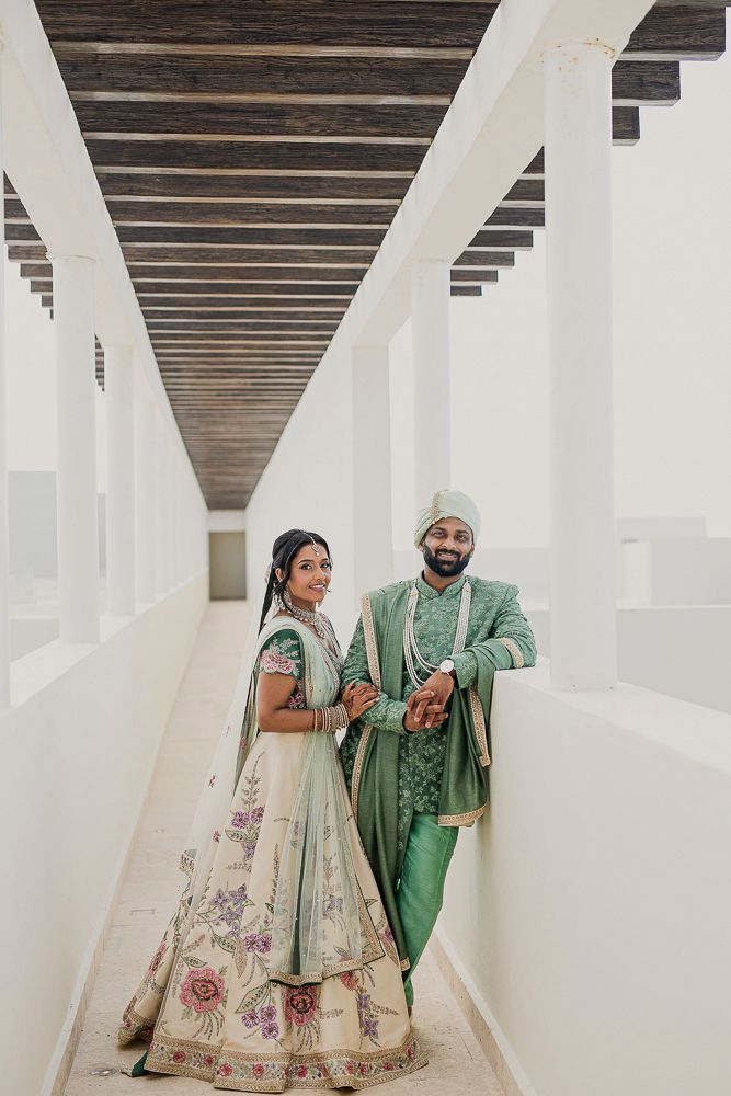 A couple in elegant green and white Indian wedding outfits pose together in a modern white interior.