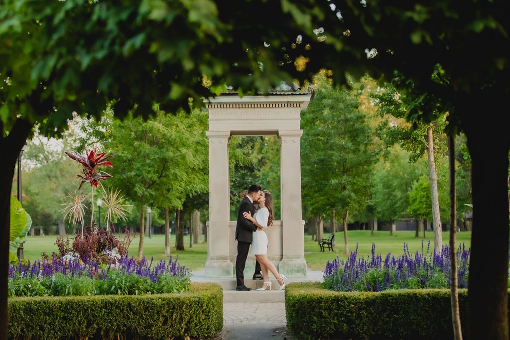 A couple shares a tender moment in a garden archway surrounded by blooming purple flowers and greenery.