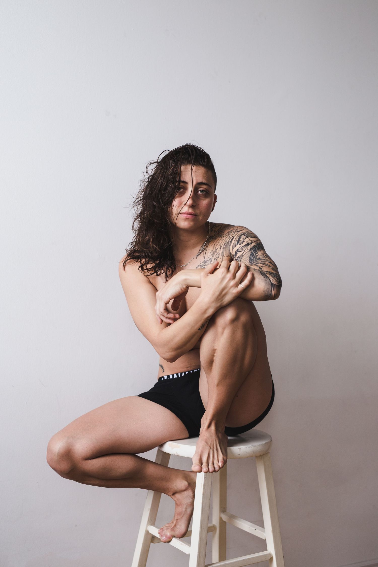 Tattooed woman sitting on white stool in black underwear, arms crossed over chest, against light gray background.