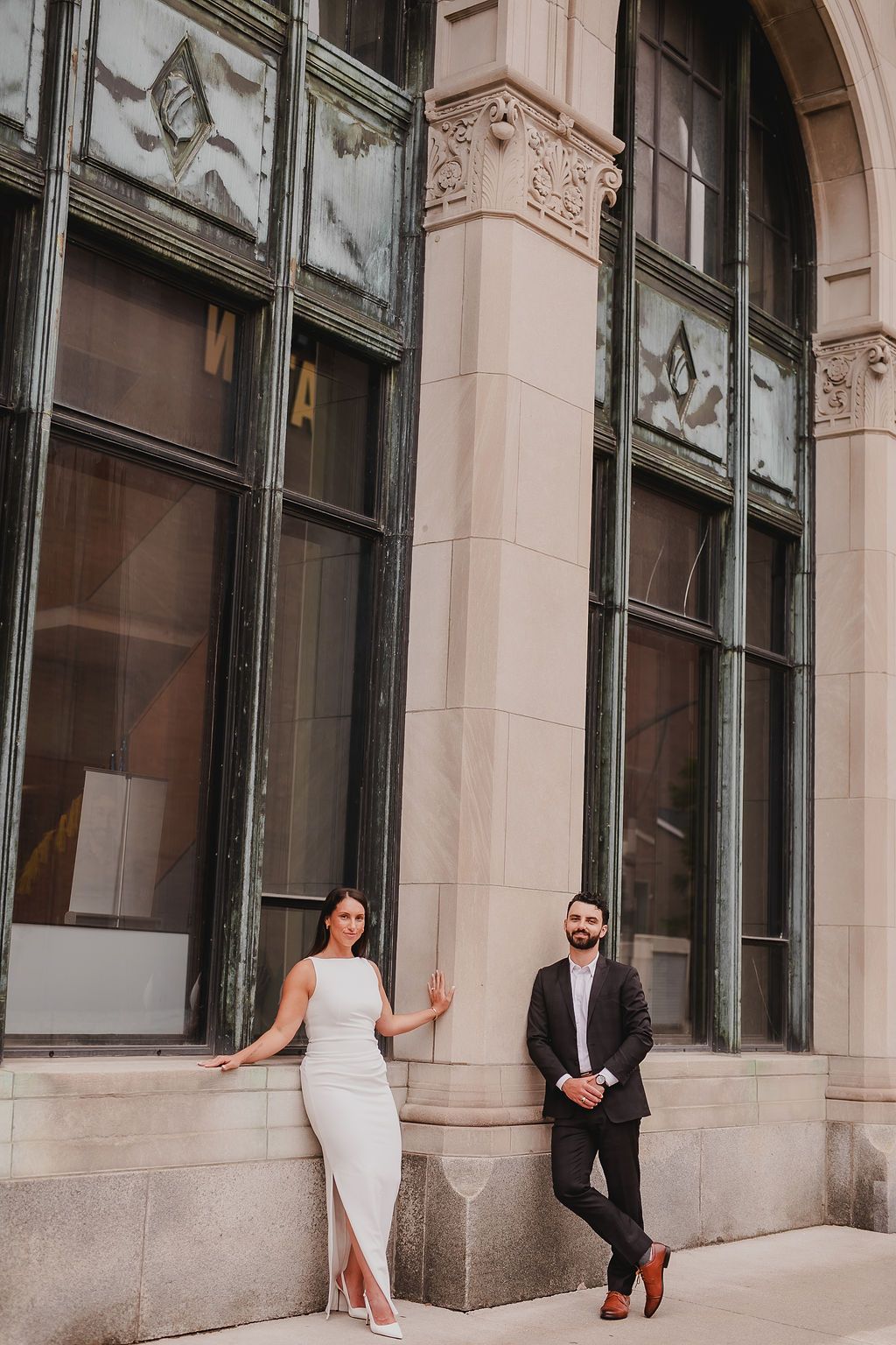 Couple in formal attire standing against ornate building exterior during golden hour.