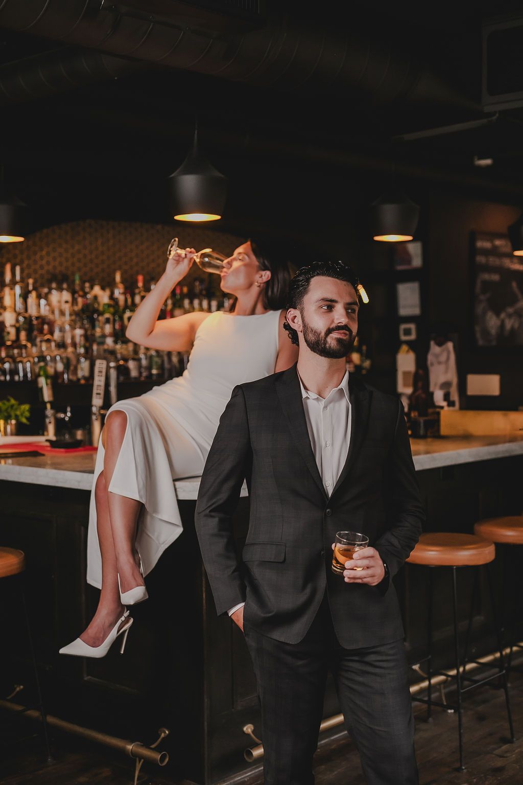 A couple shares drinks and intimate moments at a dimly lit bar with bottles displayed in the background.