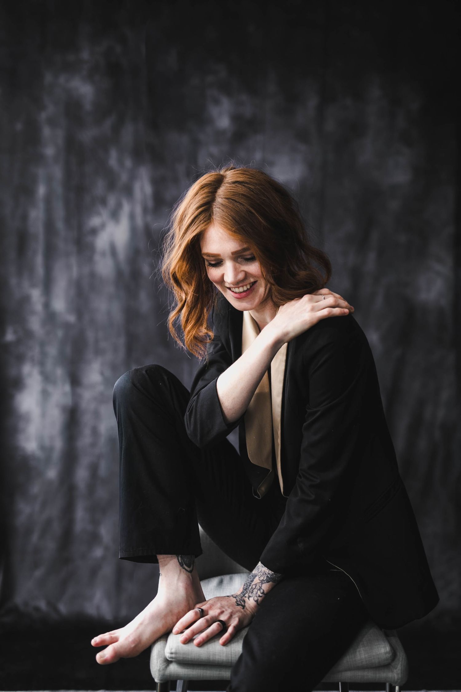 Smiling redhead woman in black suit sitting barefoot on chair, showing tattoos, against dark textured background.