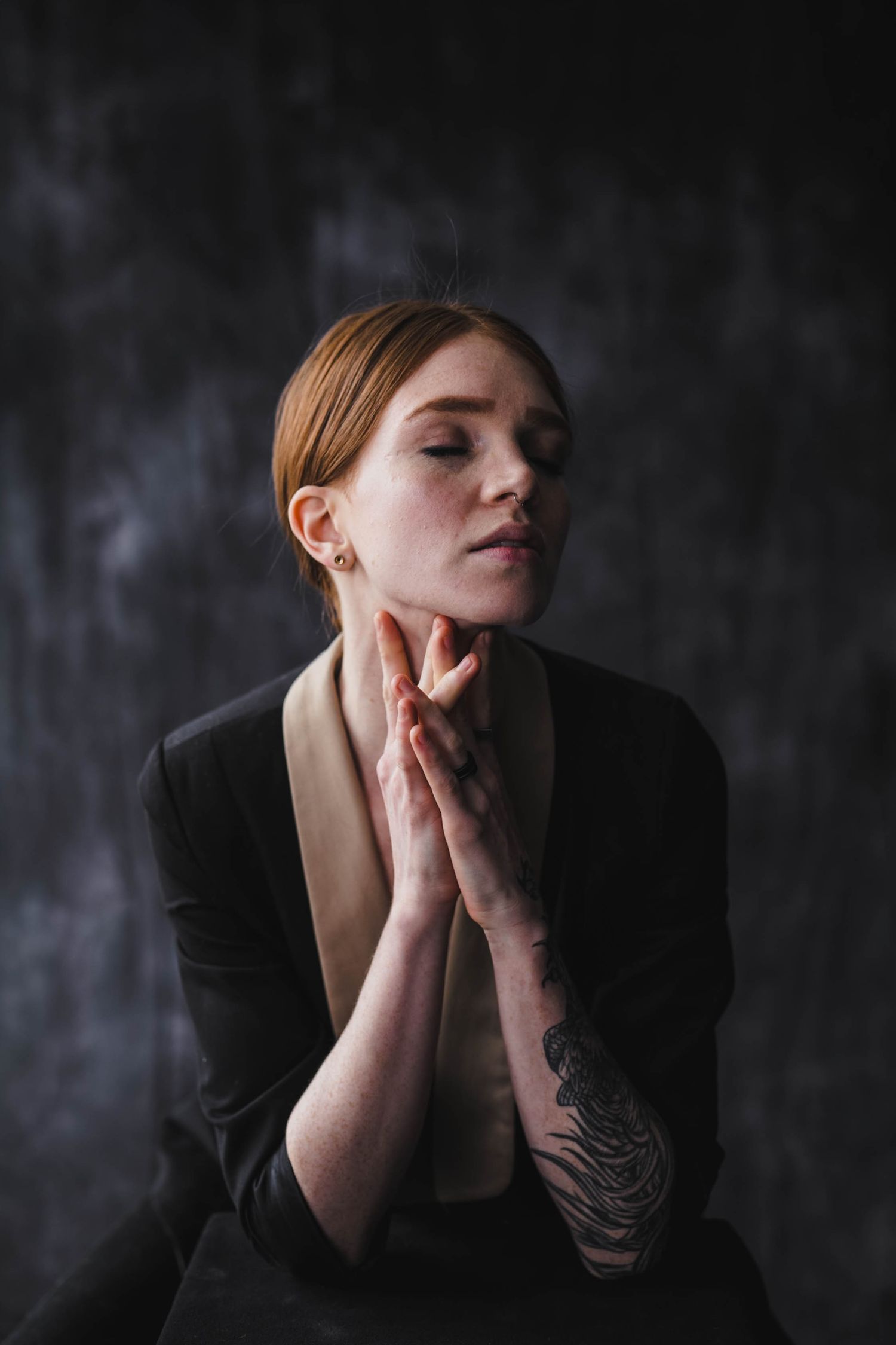 Young redhead woman with tattoo prays with hands together, eyes closed, wearing black jacket on dark background.