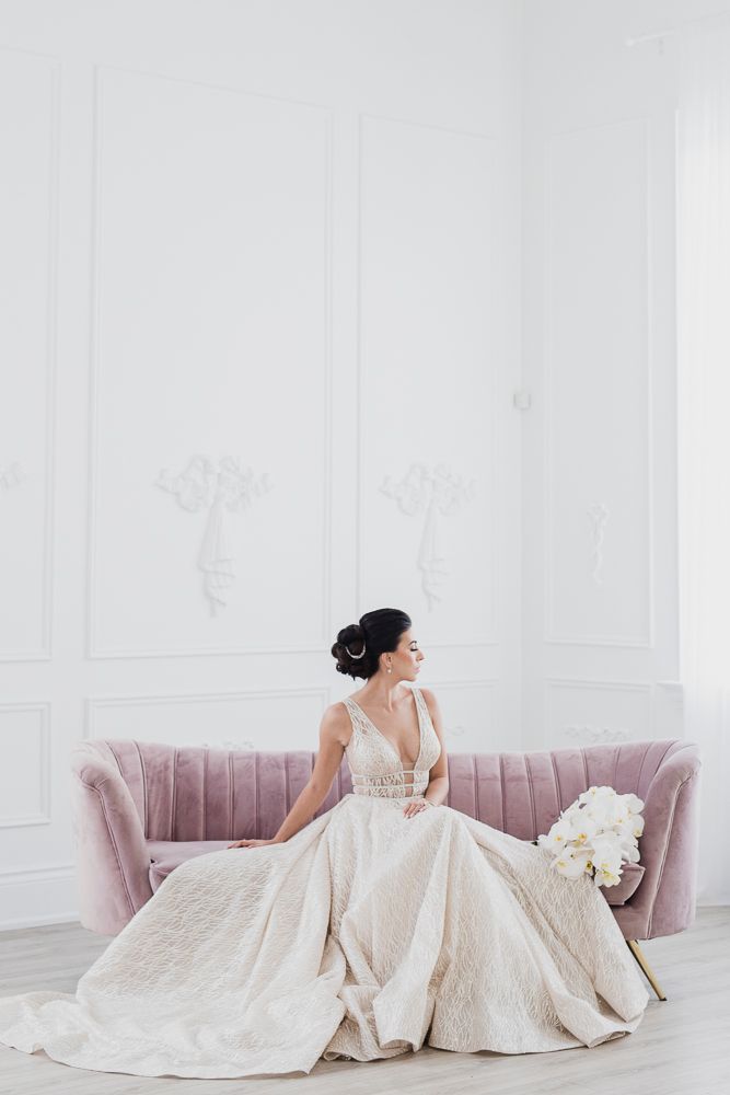 Elegant bridal portrait on pink velvet sofa showing detailed white wedding gown in bright studio setting.