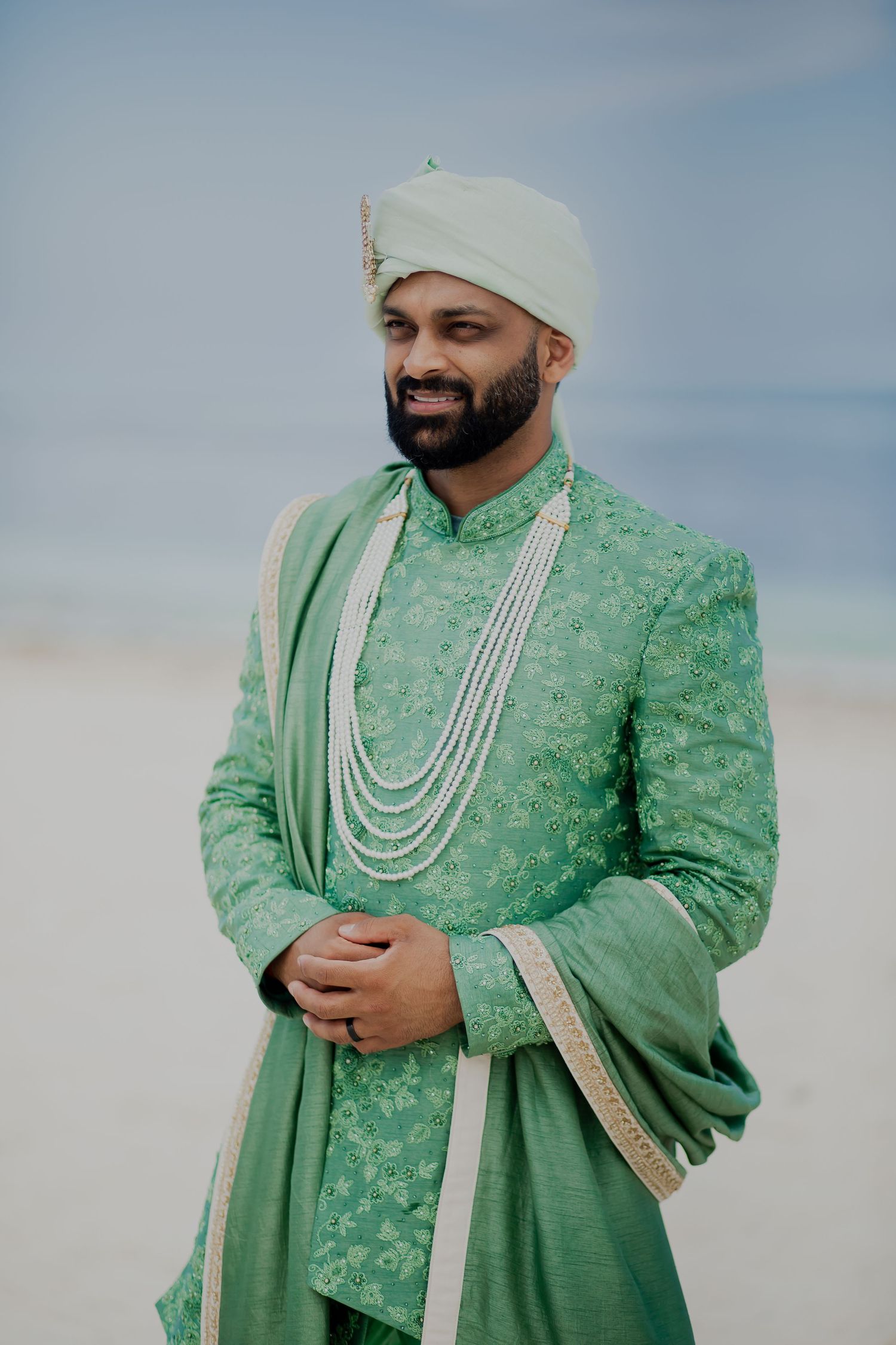 Groom in mint green sherwani with pearl necklaces poses on beach.