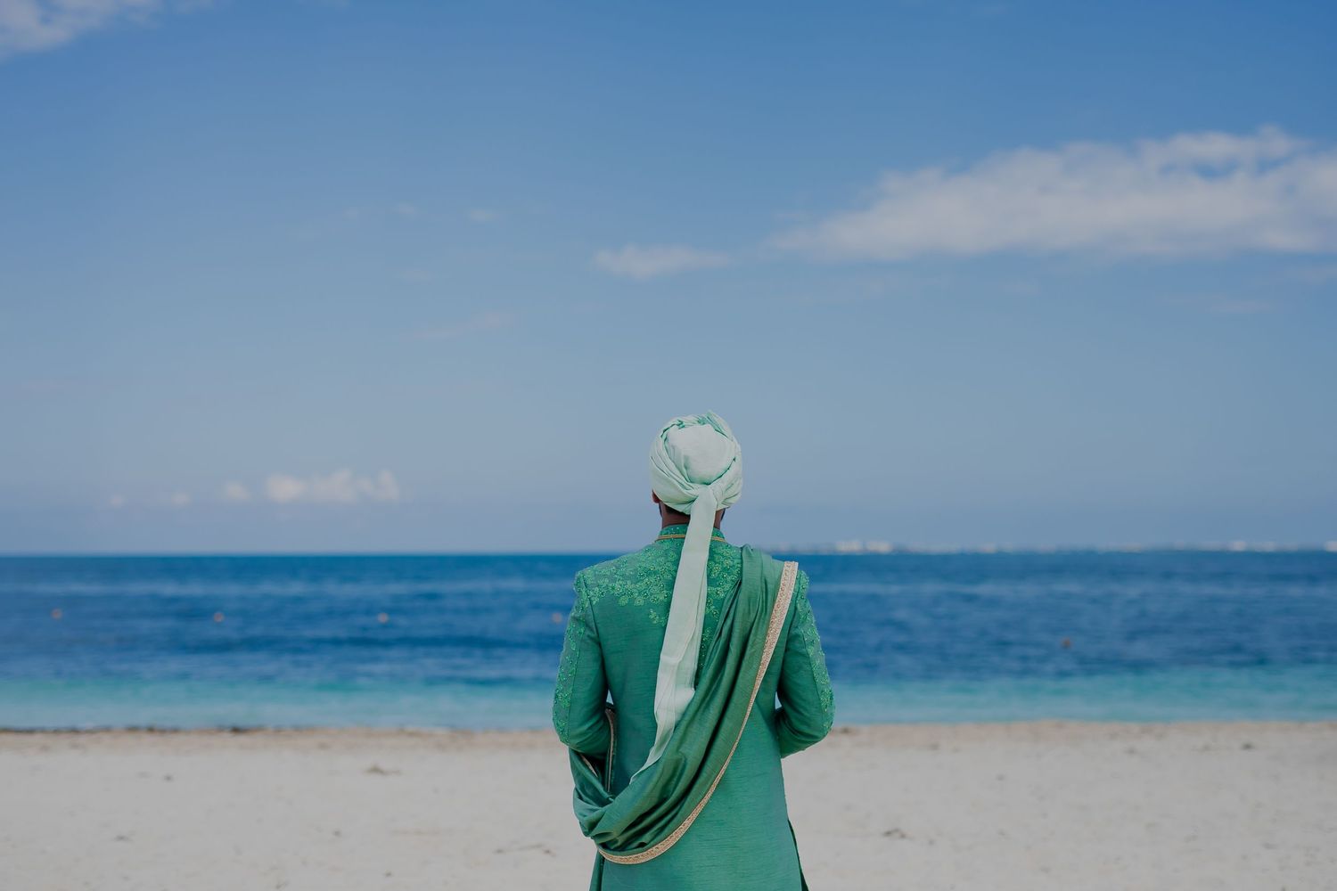 A person in green traditional attire stands on a beach overlooking turquoise ocean waters.