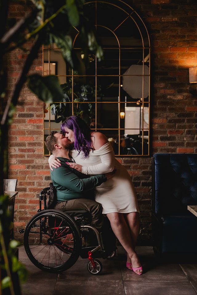 A couple shares an intimate indoor moment with one partner seated in a wheelchair against a rustic brick wall.