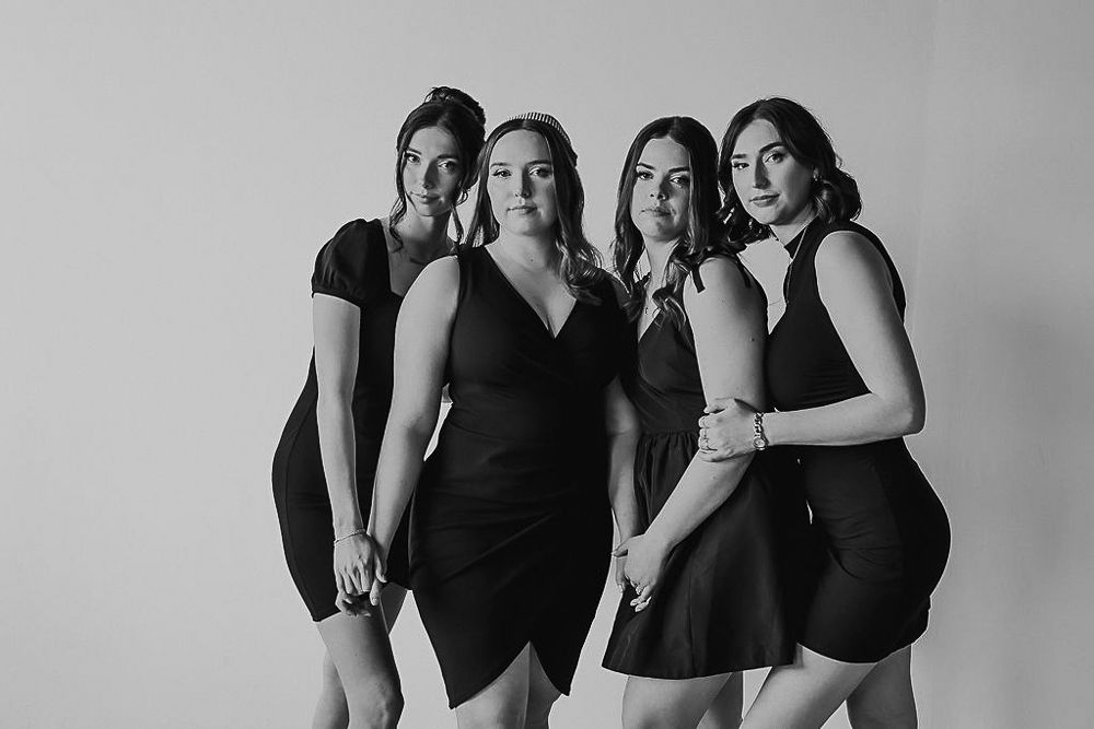 Four women in black dresses pose together against a light background in a black and white photo.