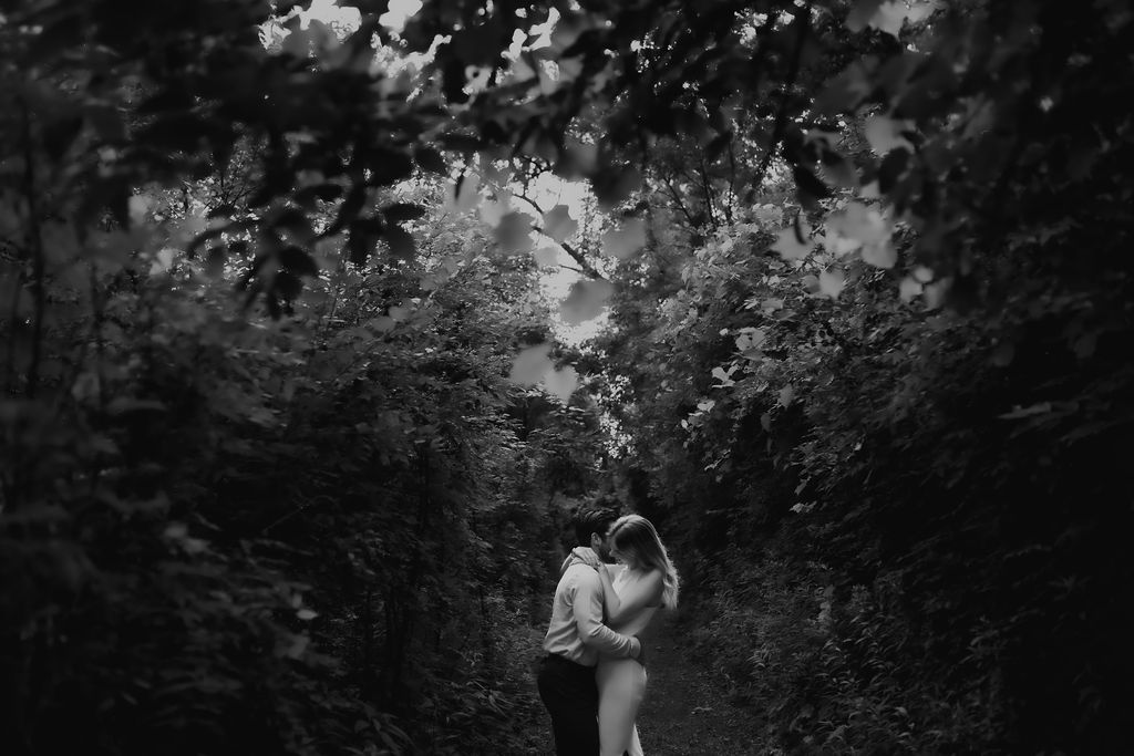 A romantic black and white photo of a couple embracing on a wooded path surrounded by trees.