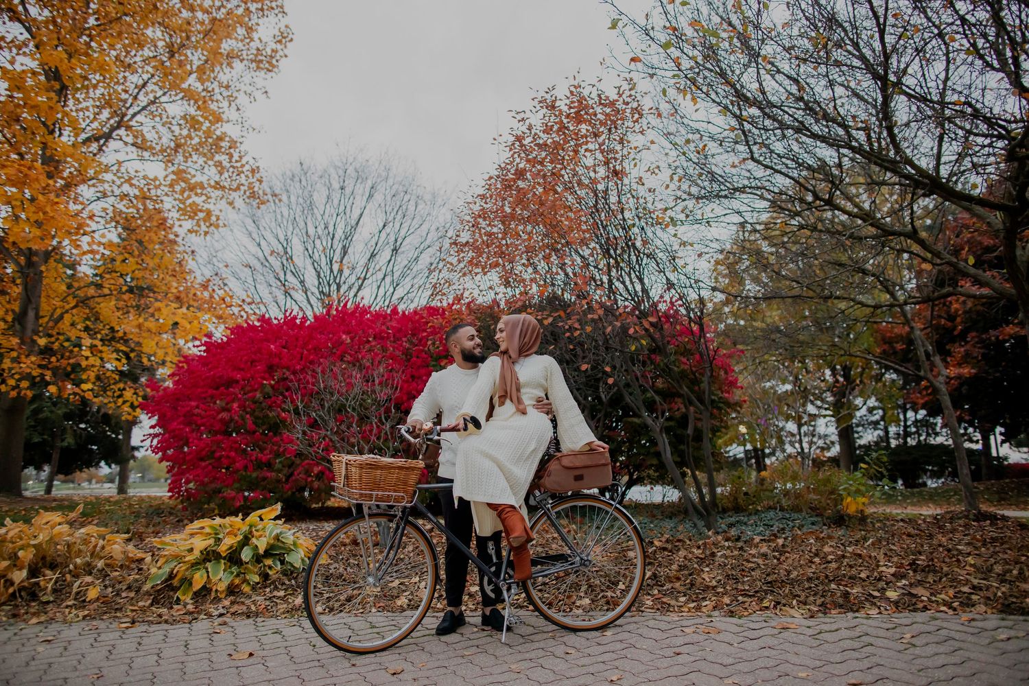 A couple sits together on a park bench surrounded by vibrant autumn foliage and bright red bushes.