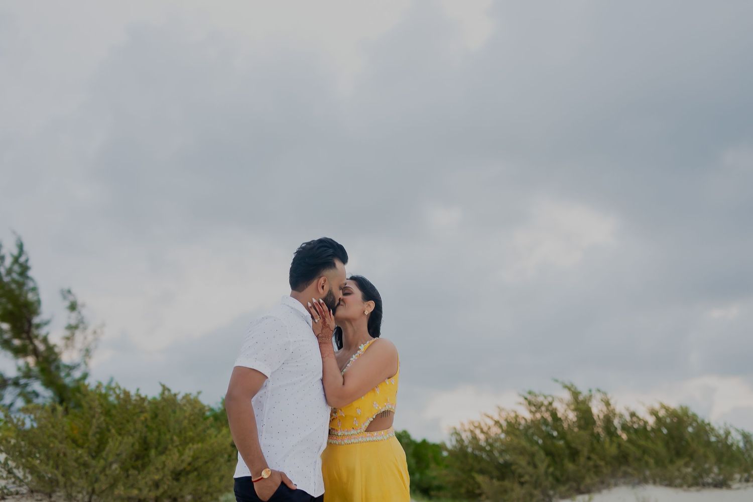 A couple embraces against a cloudy sky with greenery in the background.