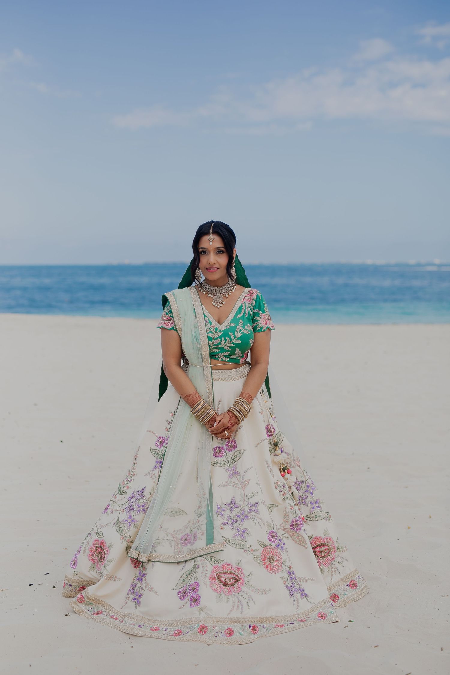 Bride wearing floral lehenga stands on white sandy beach against turquoise ocean backdrop.