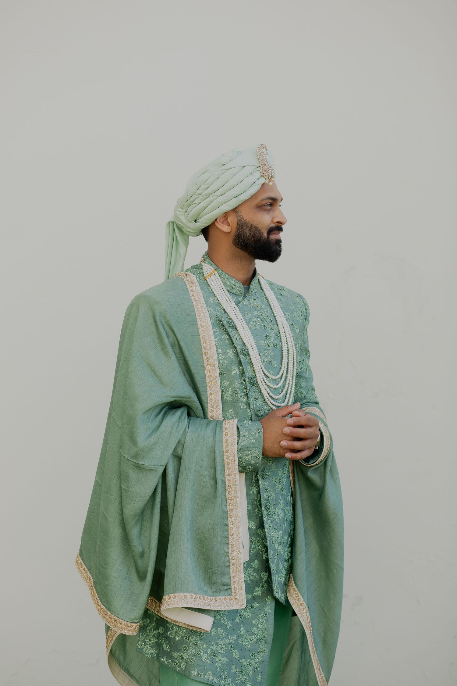 Man in mint green sherwani and turban with pearl necklaces against light background.