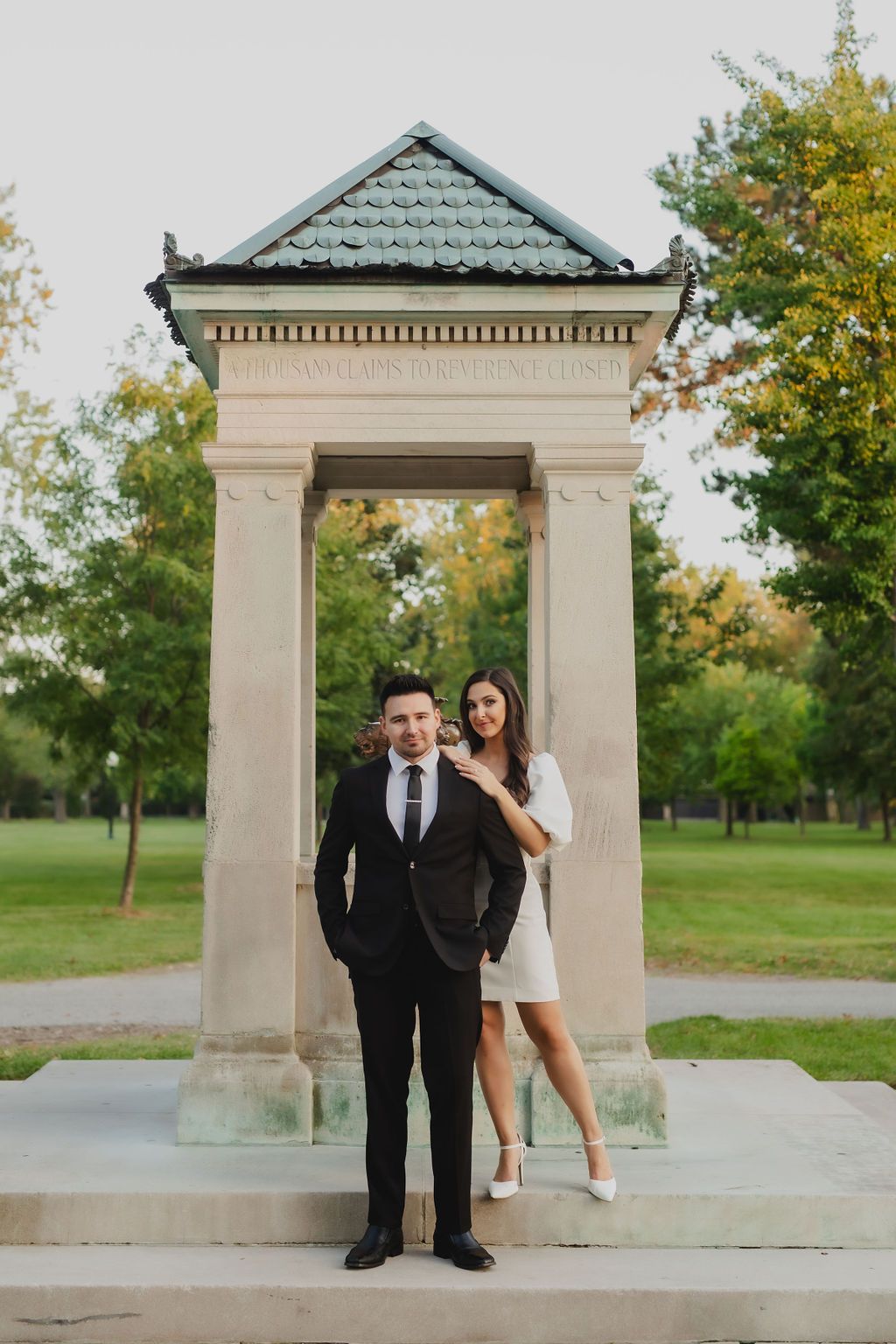 Elegant couple poses at stone archway in park setting during sunset photo session.
