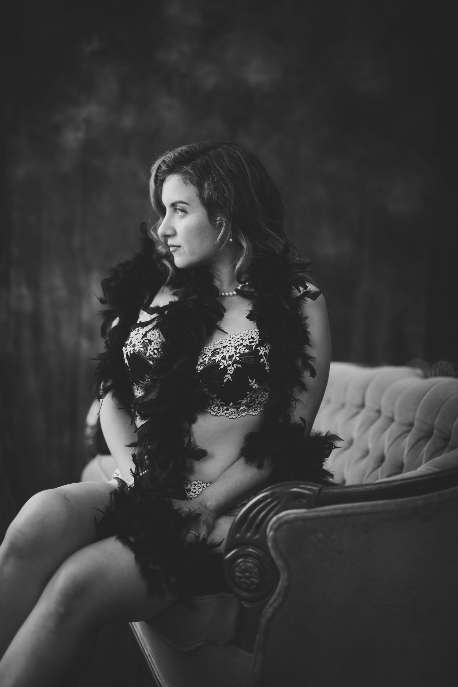 Black and white boudoir photo of a woman in lace lingerie and feather boa seated on a vintage tufted chair.
