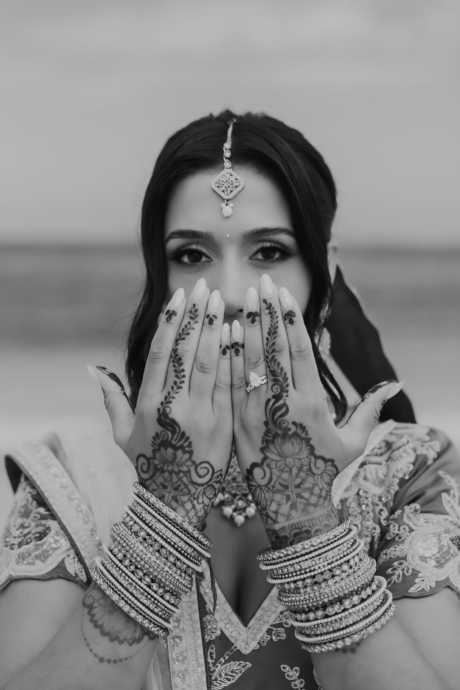 Black and white photo showing decorated hands with henna and bangles covering face.