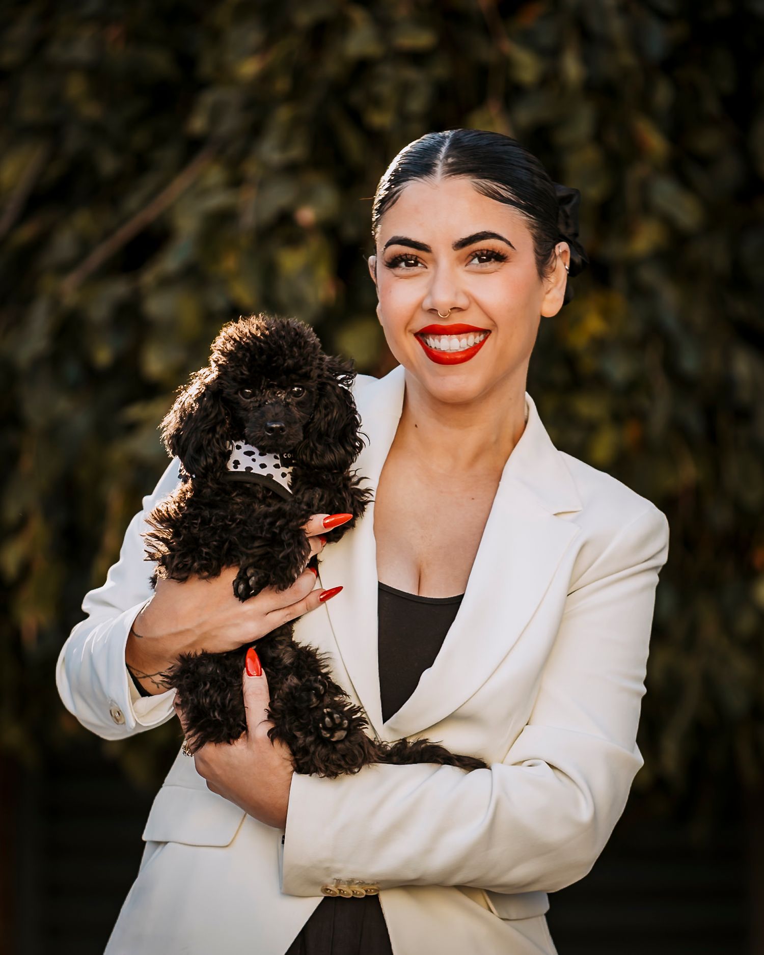 A woman in a white blazer lovingly cuddles with a small black poodle dog in a series of outdoor portraits.