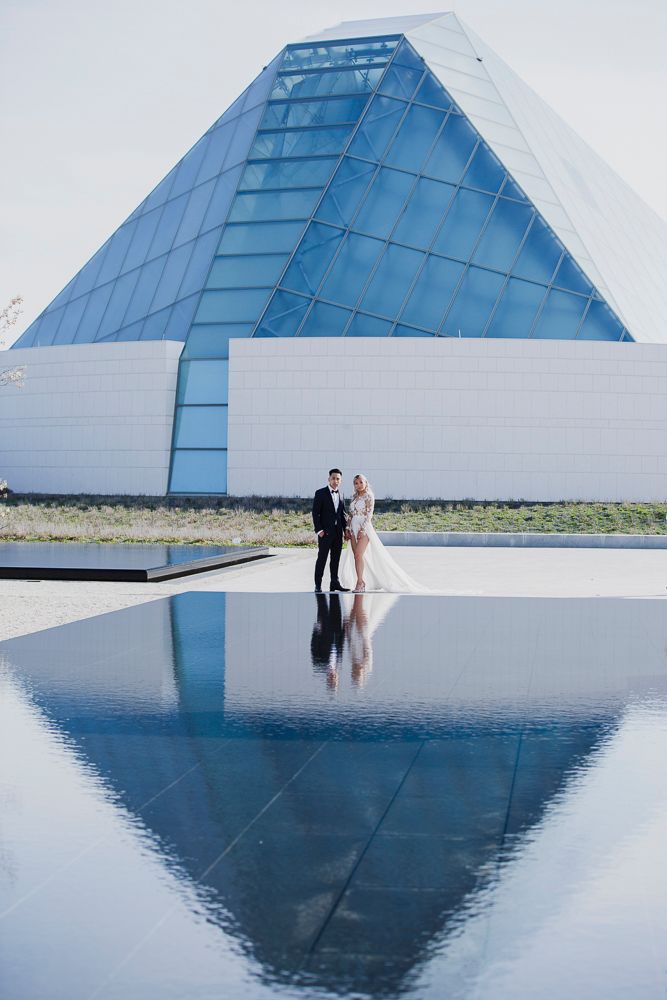 Couple stands by reflective pool beside modern geometric blue and white pyramid architecture in minimalist setting.