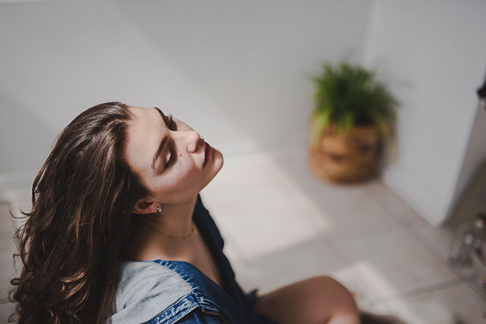Natural light portrait showing relaxed pose near window with indoor plants in background.
