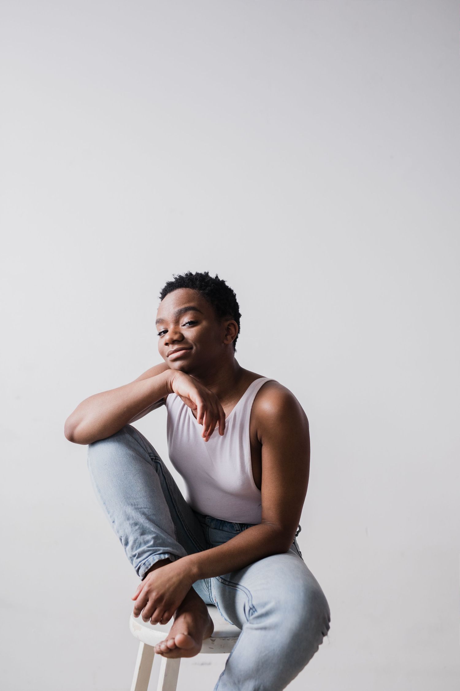 Young Black person with short hair sits on a stool in jeans and tank top, resting chin on hand against gray background.