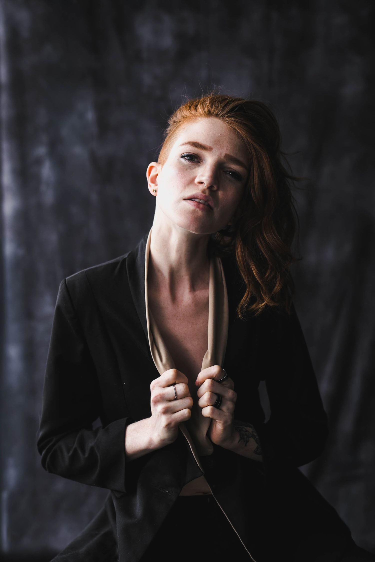 A redhead woman in a black blazer poses dramatically against a dark textured background in a moody portrait.
