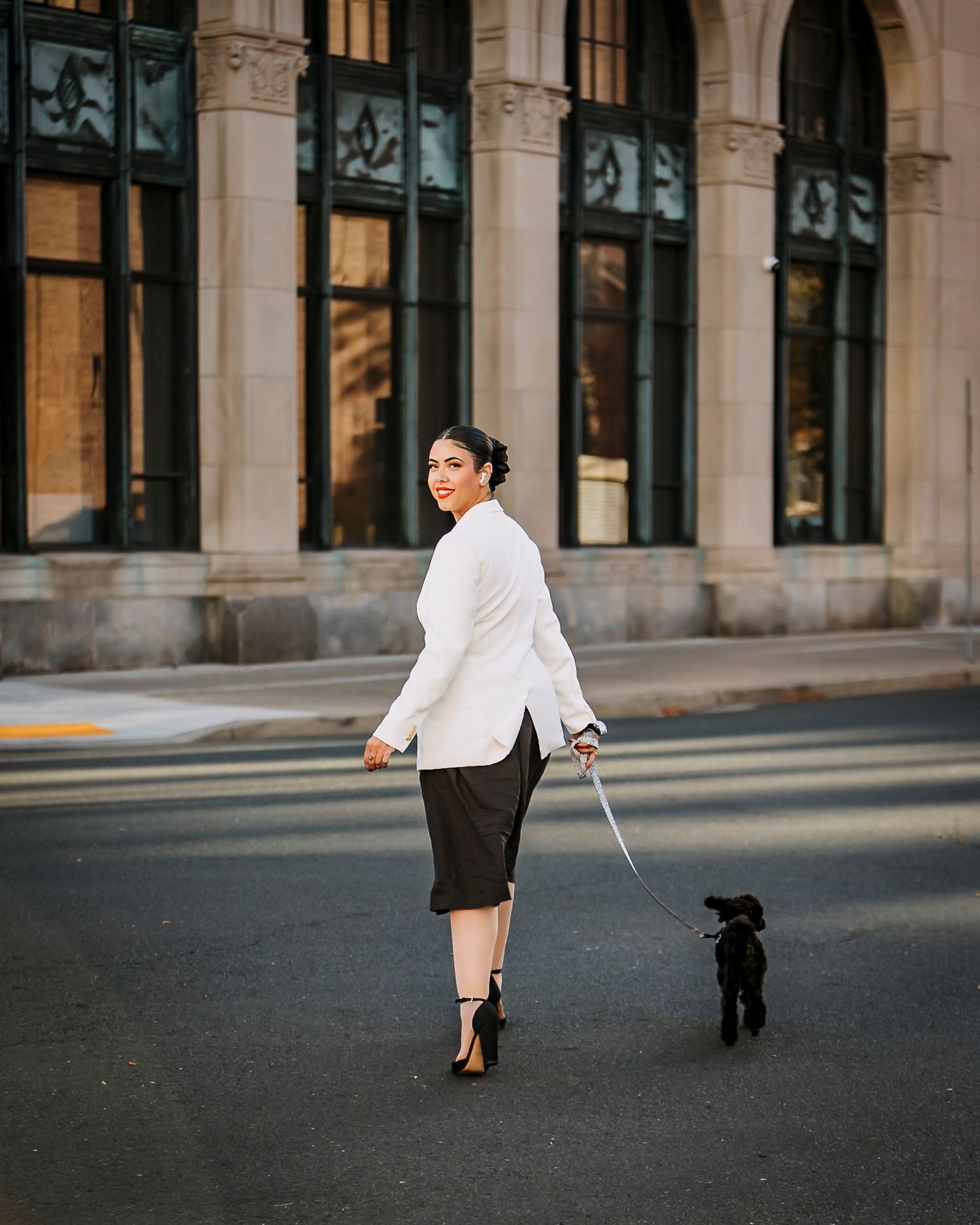Woman in white blazer and black skirt walks small black dog on leash across urban street intersection.