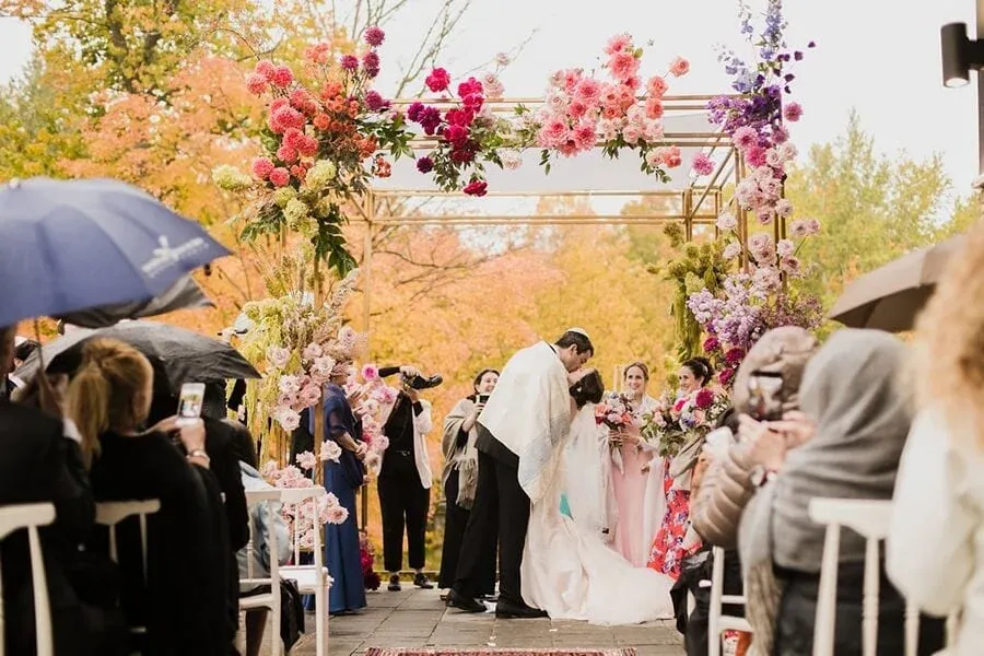 Pink flower petals float through the air during an autumn outdoor wedding ceremony beneath golden trees.