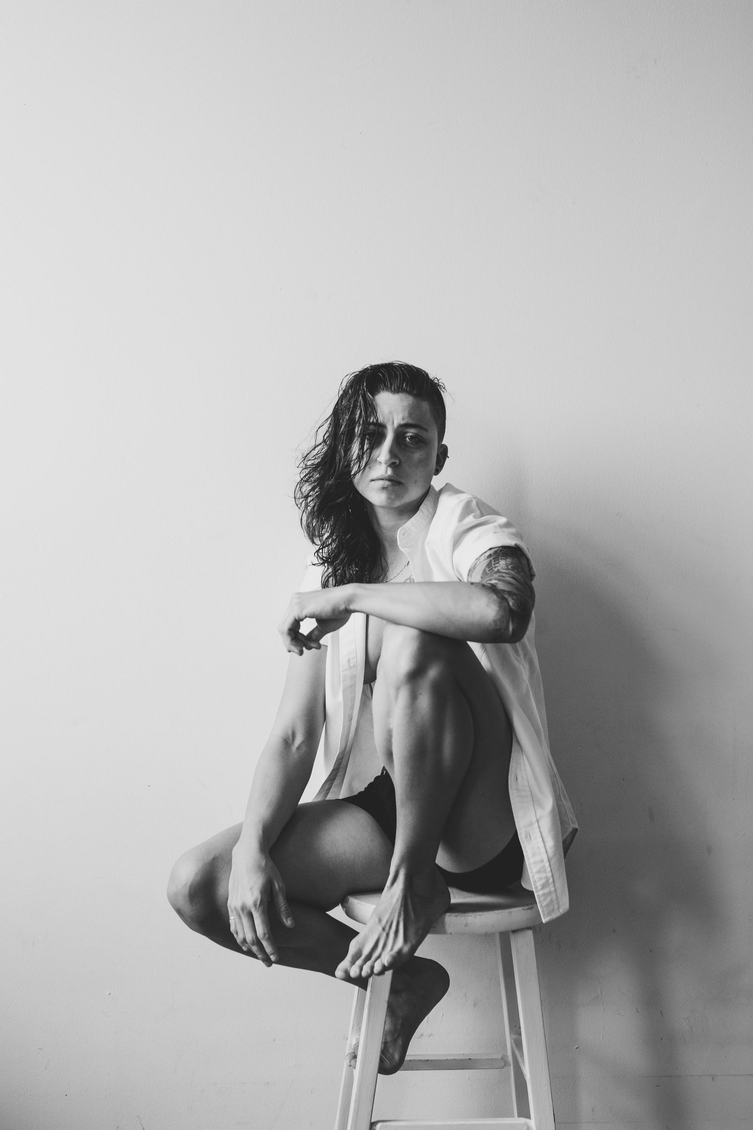 Black and white photo of a young woman sitting cross-legged on a stool, wearing an open white shirt, gazing at the camera.