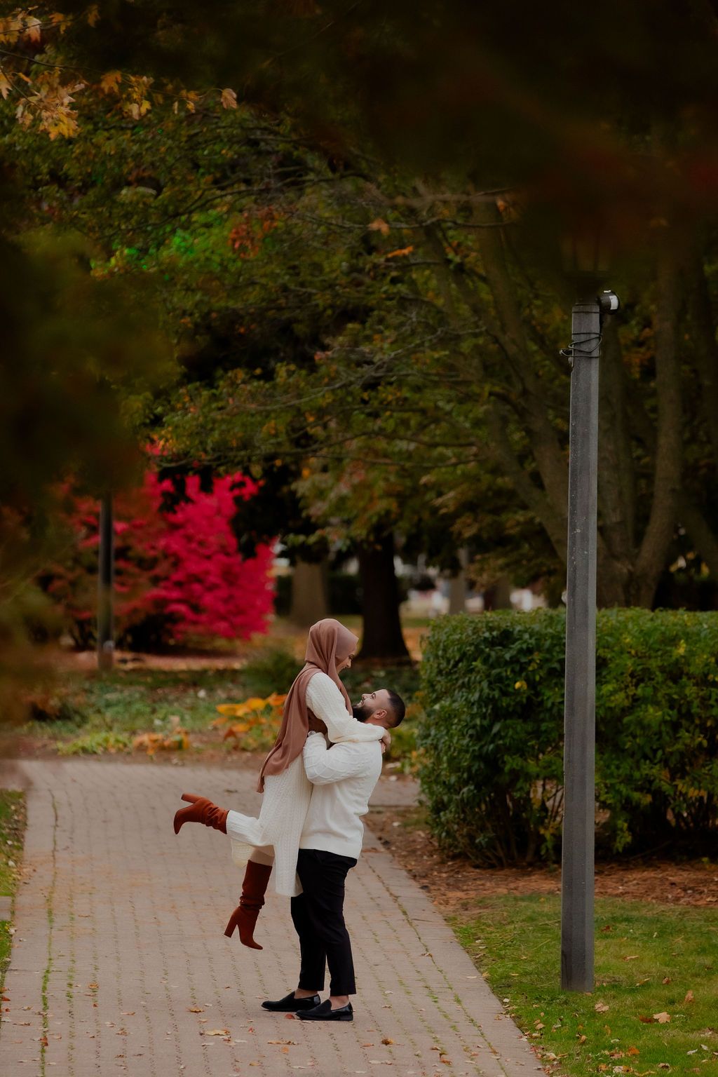 A couple embraces on a brick pathway surrounded by autumn foliage and trees with vibrant red leaves in the background.