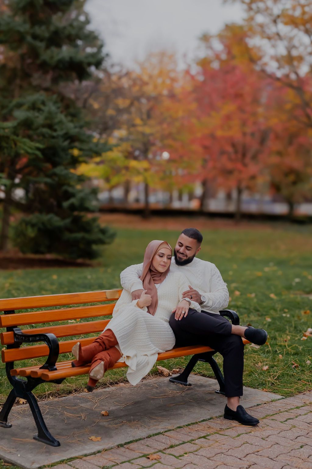 A couple relaxes together on a wooden park bench surrounded by colorful fall foliage and trees.