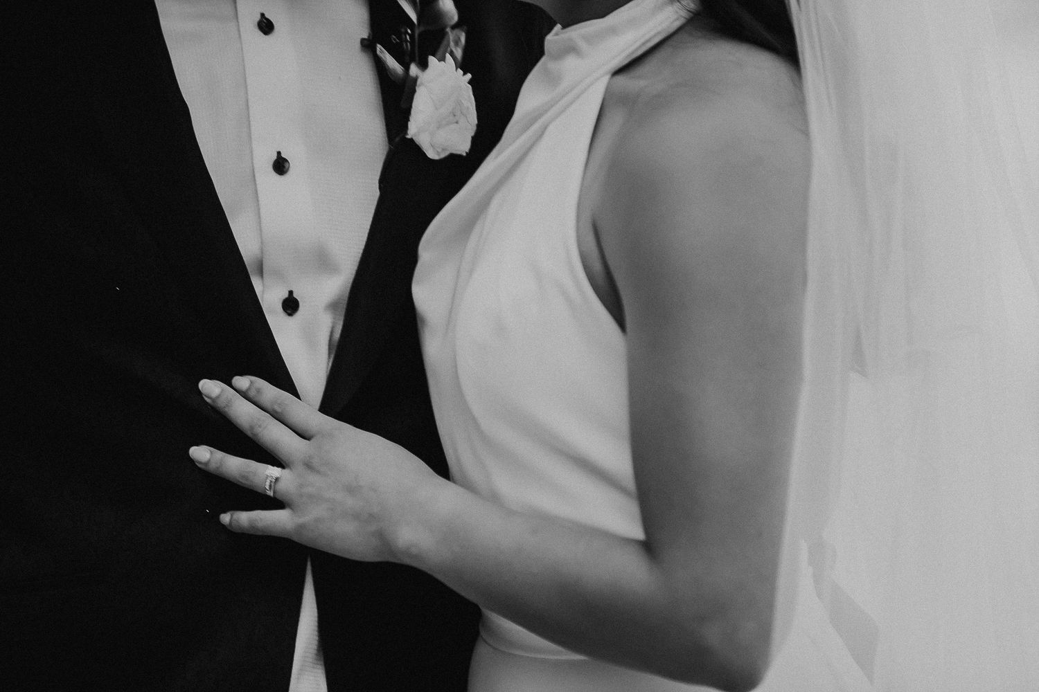 Black and white wedding photograph showing hands with rings and white formal attire details.