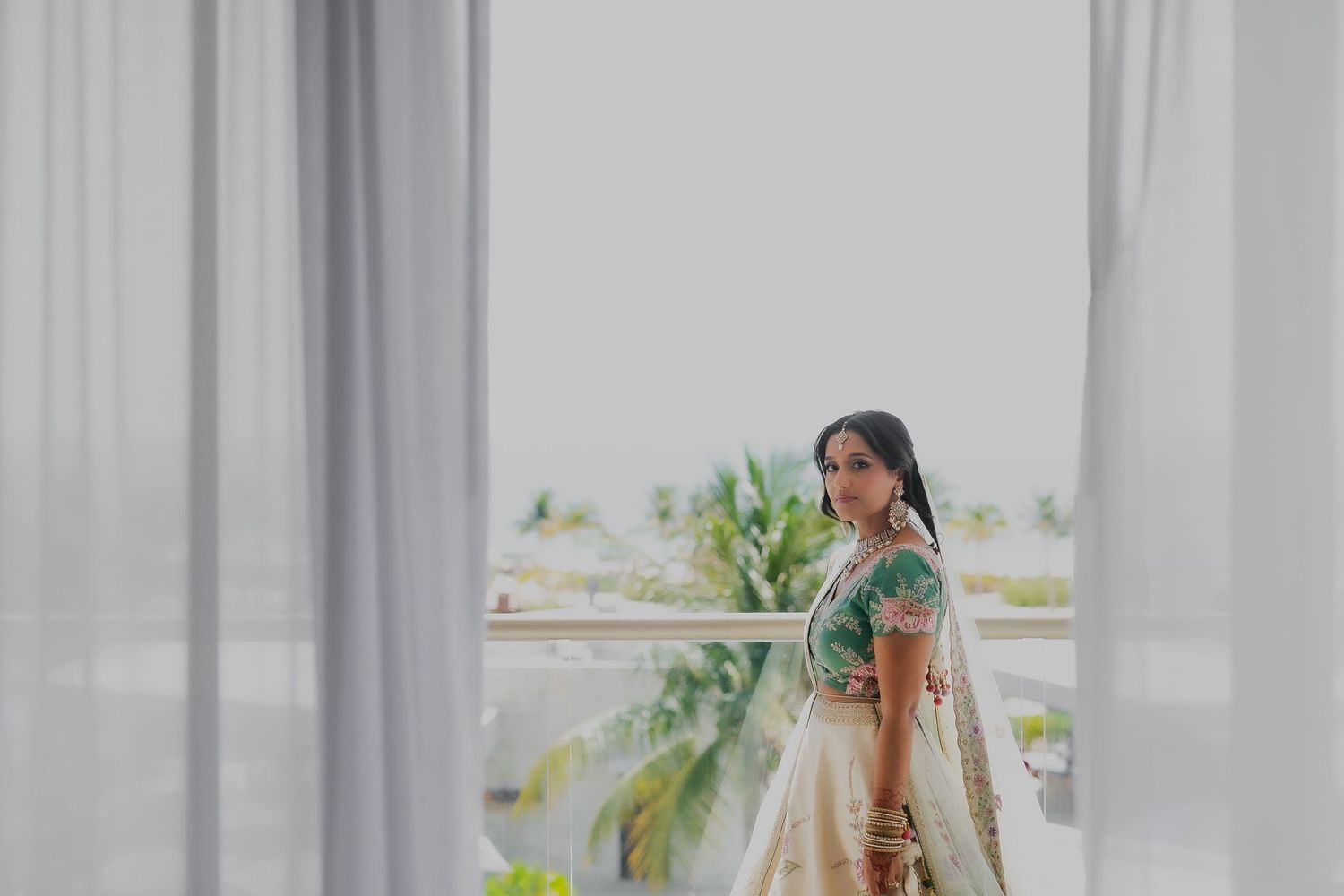 Bride in mint green traditional dress standing by curtained window with tropical view.