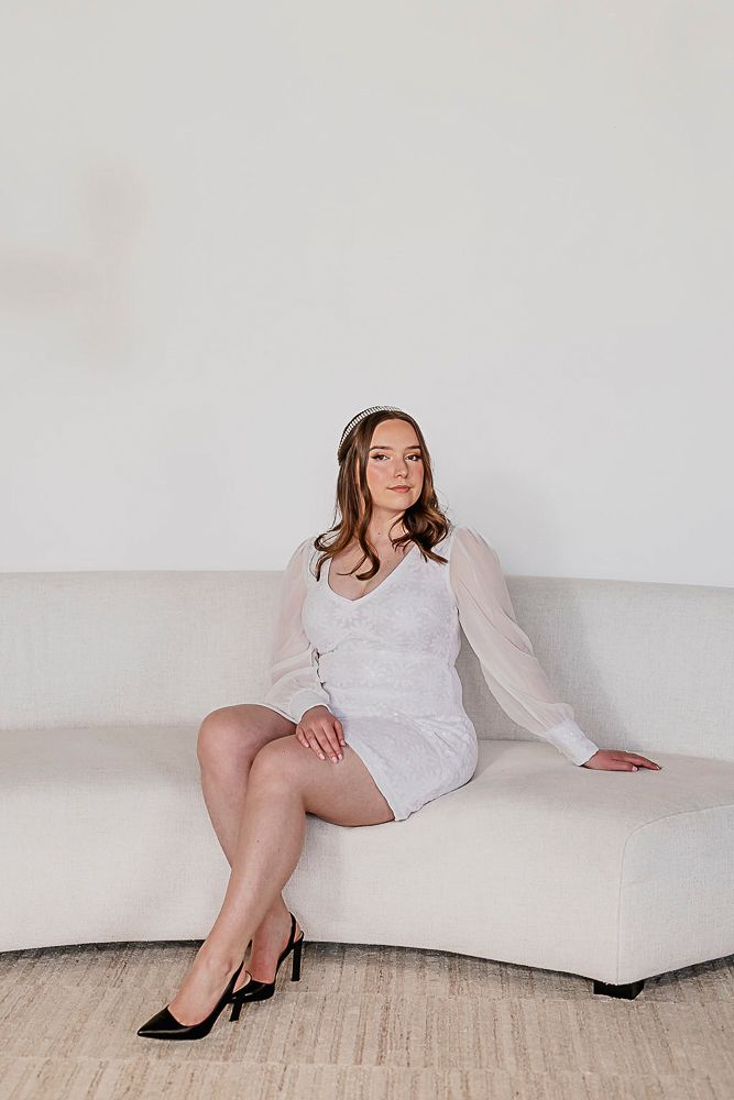 A person in a white long-sleeved dress poses elegantly on a light-colored couch with black high heels.
