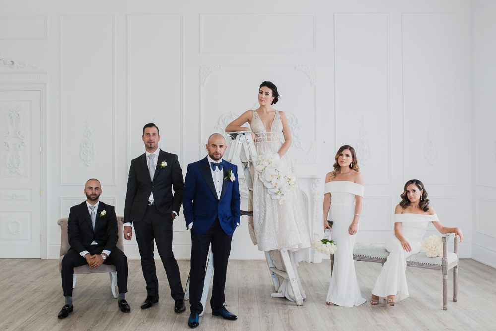 Modern wedding party in formal attire poses elegantly against white wall in minimalist studio setting.