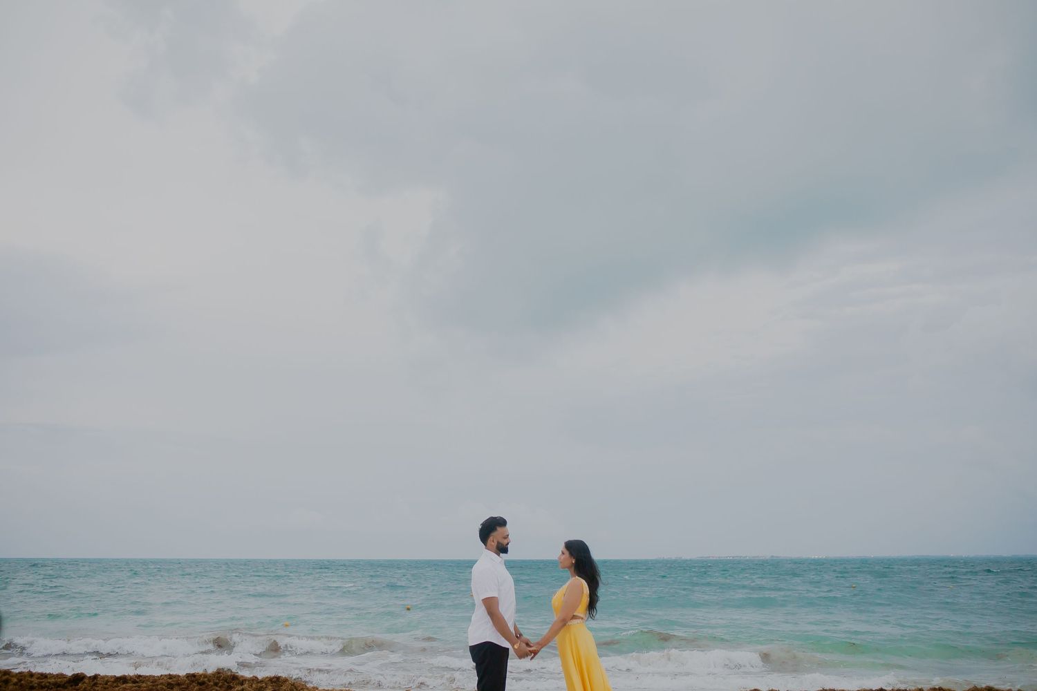 A couple stands together on a beach with turquoise ocean waters in the background.