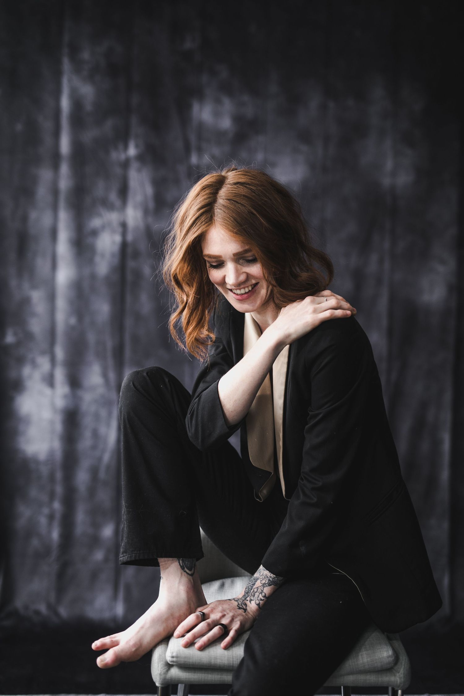Smiling redhead woman in black suit sitting on chair with tattoos on hands against dark background.