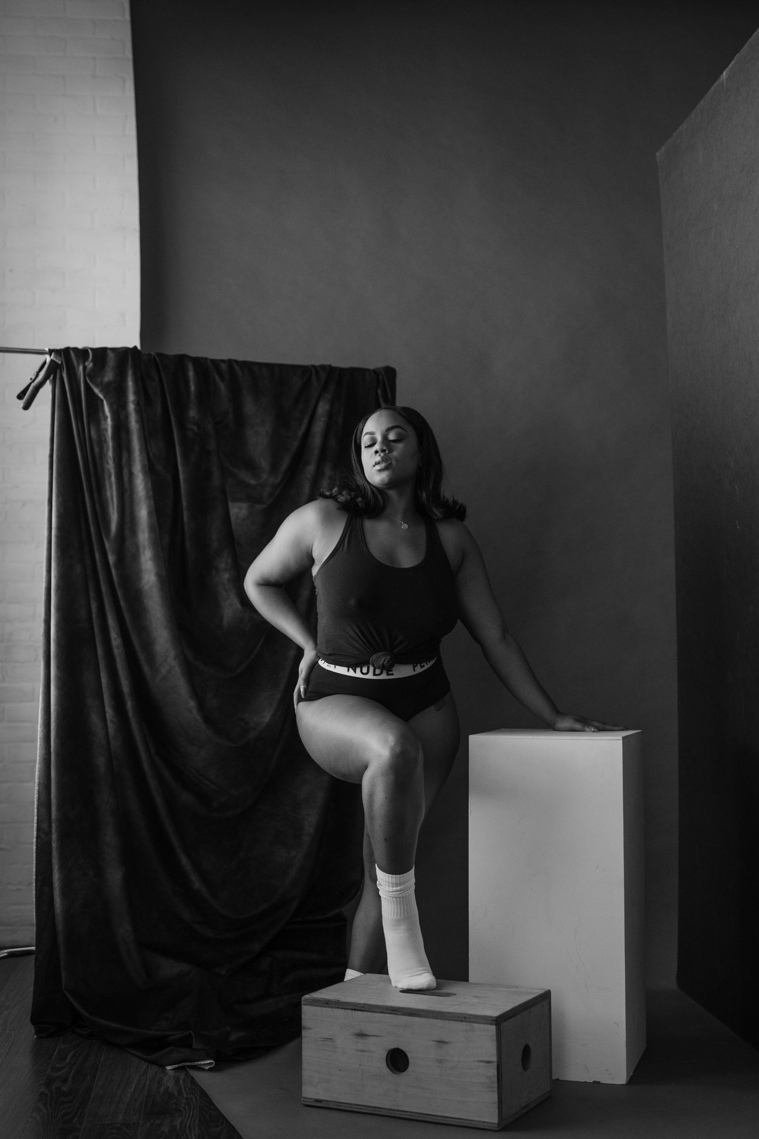 Black and white studio photo of a plus-size woman in athletic wear posing confidently on a wooden box.