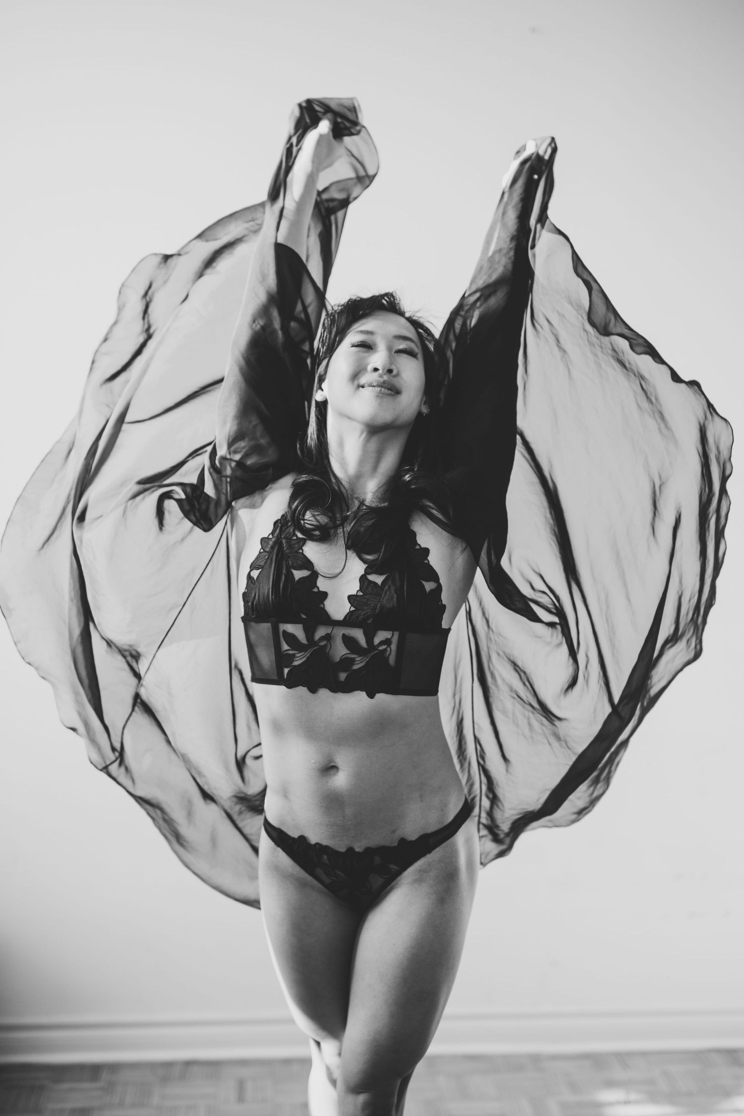 Black and white photo of a woman in lingerie gracefully holding sheer fabric above her head like wings.