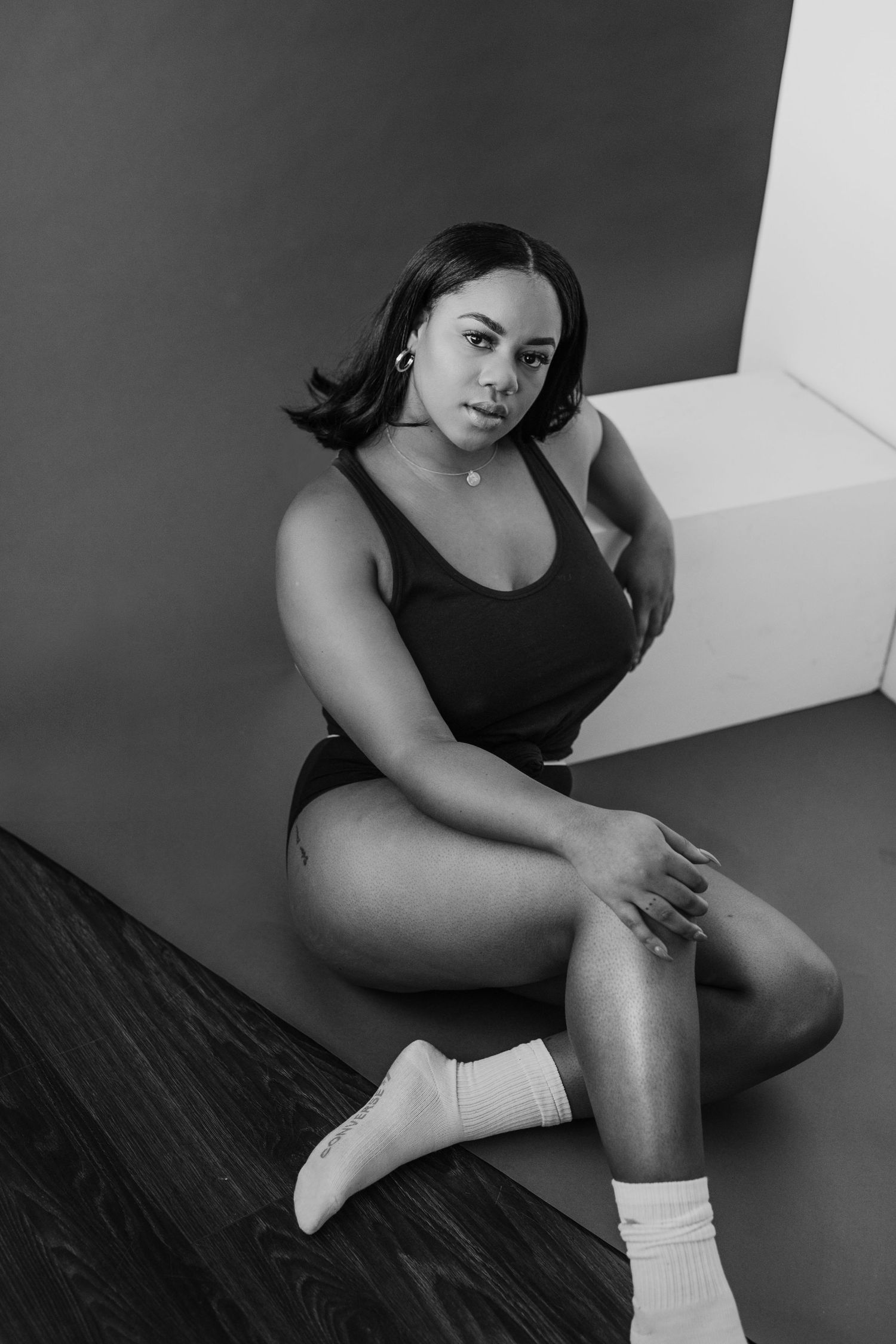 Black and white photo of a young woman in a black bodysuit and white socks sitting on a dark wood floor.