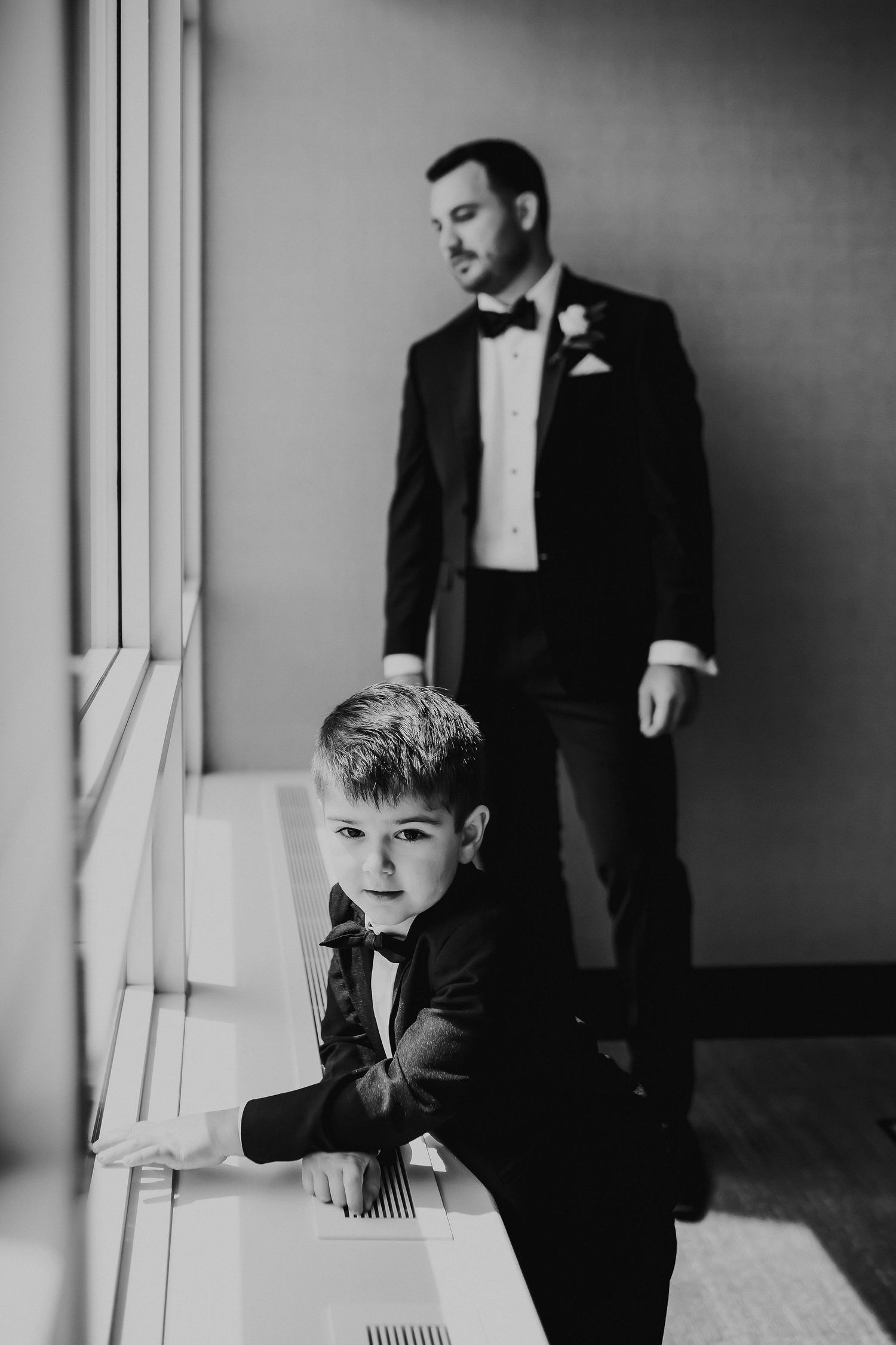 Young boy in formal suit sitting pensively by window in moody black and white setting.