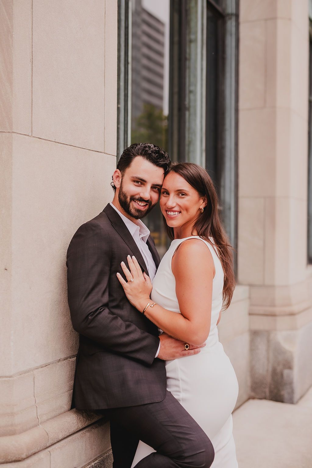 A couple embraces and laughs together against a light stone building exterior.
